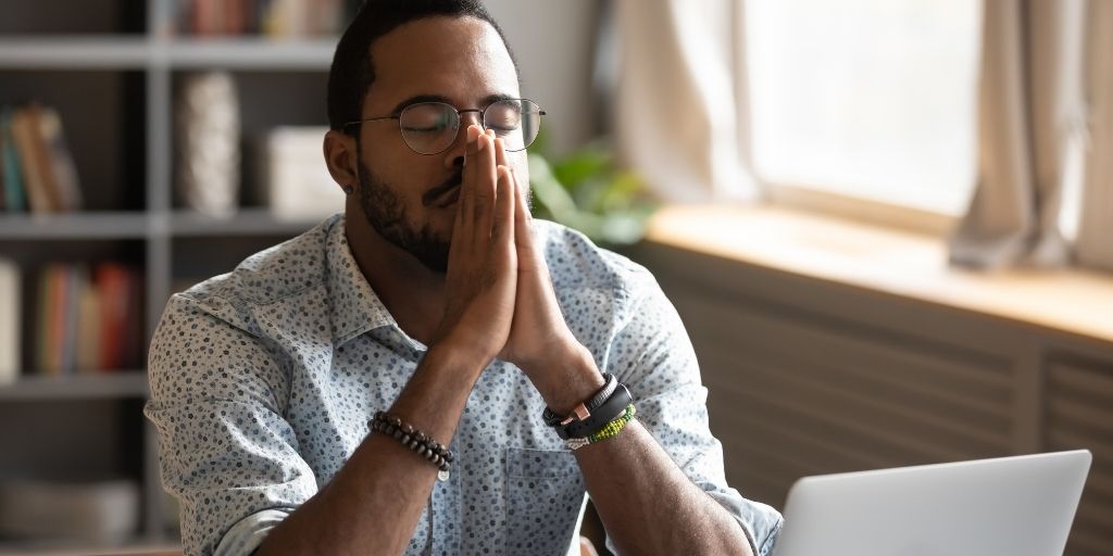 african american male struggling with mental health. appears with eyes closed, elbows resting on desktop and hands held palm to palm in front of face as if in prayer