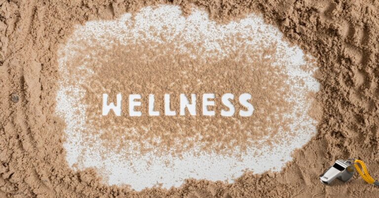wellness written in sand, coaches whistle laying on sand in foreground